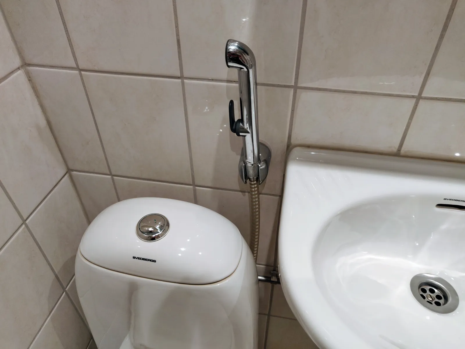 Douchette WC chromée fixée au mur carrelé, entre des toilettes blanches et un petit lavabo