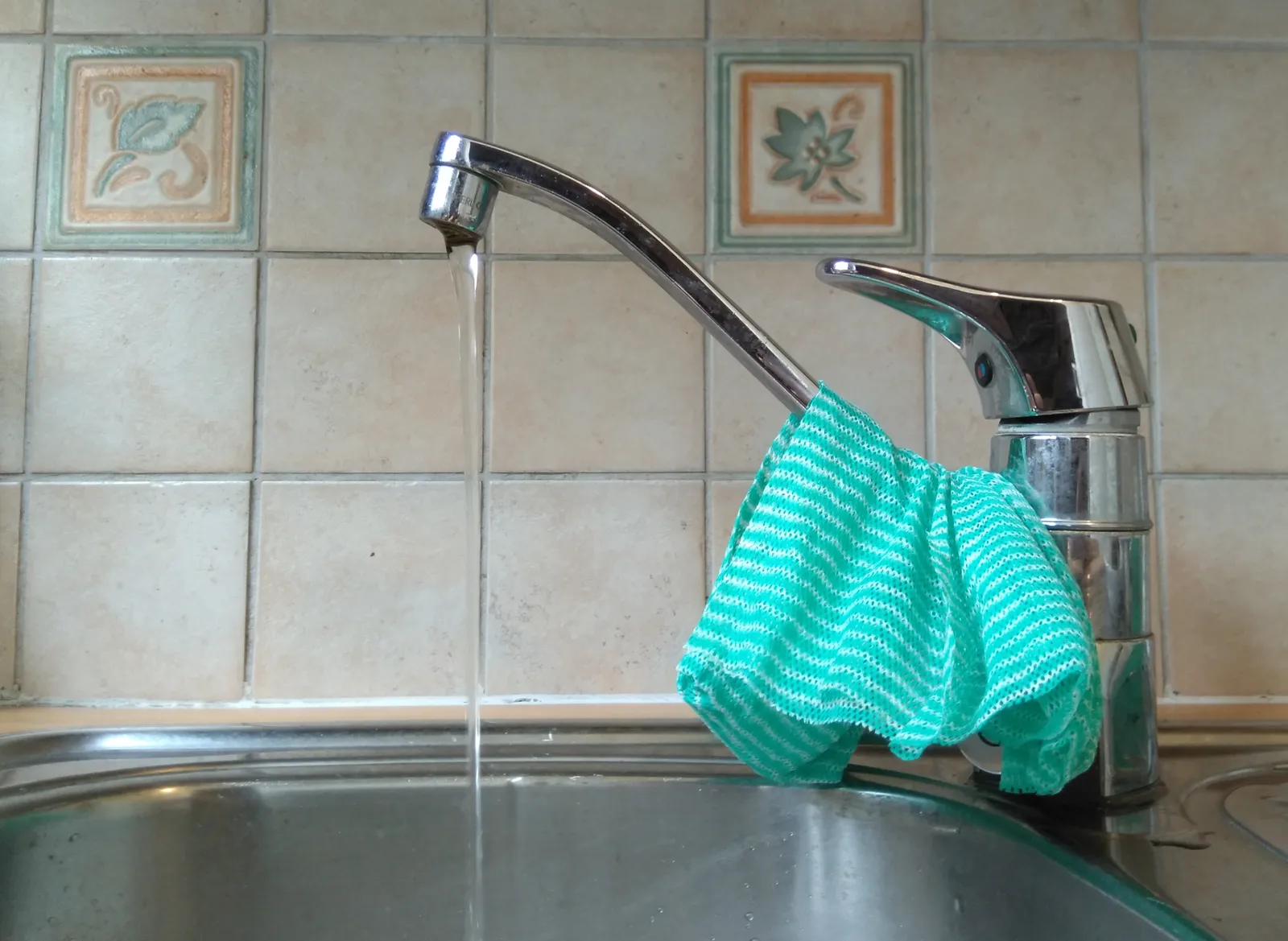 Filet d'eau qui coule d'un robinet chromé dans un évier en inox, devant un mur carrelé