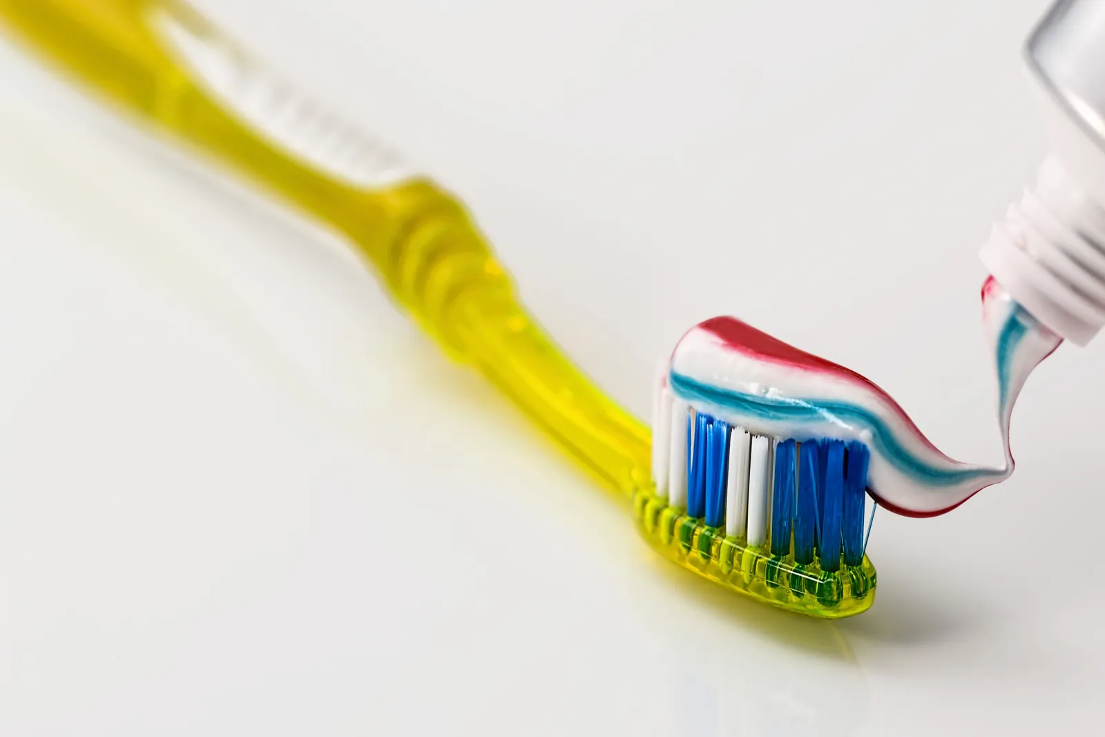 Brosse à dents jaune aux poils bleus et blancs sur laquelle on dépose du dentifrice rayé rouge et bleu, sur fond blanc