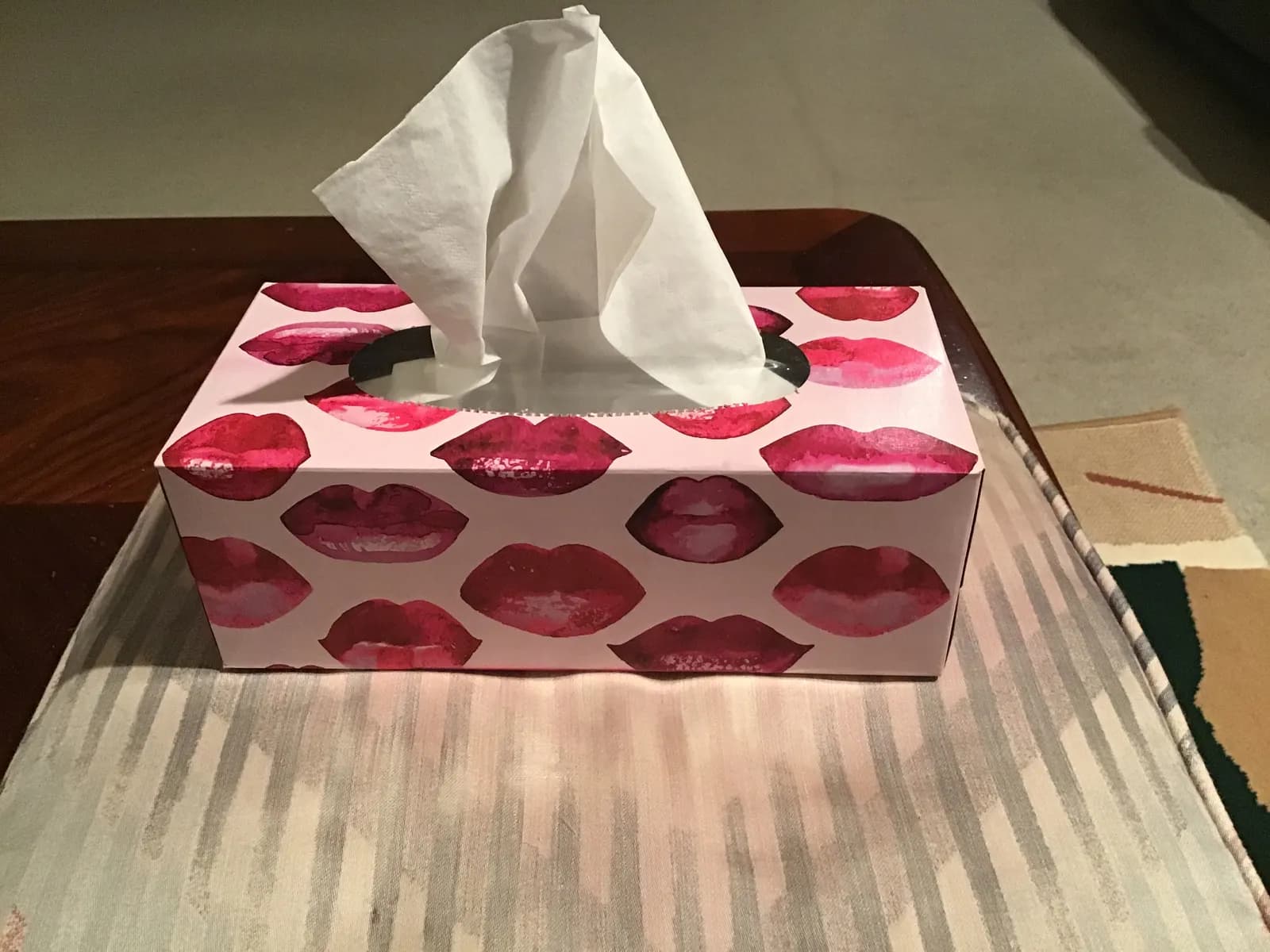 Boîte de mouchoirs en papier décorée de motifs de lèvres, un mouchoir dépassant de l'ouverture, posée sur un accoudoir