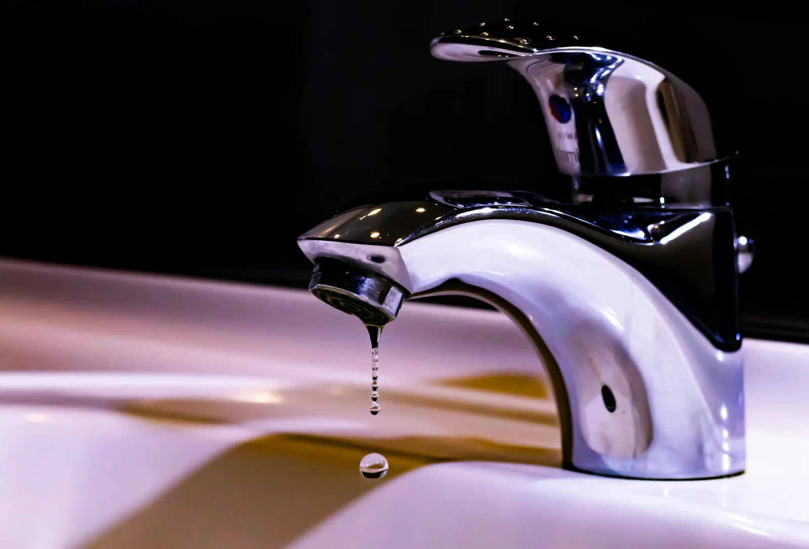 Robinet chromé d'où tombe une goutte d'eau sur un lavabo blanc