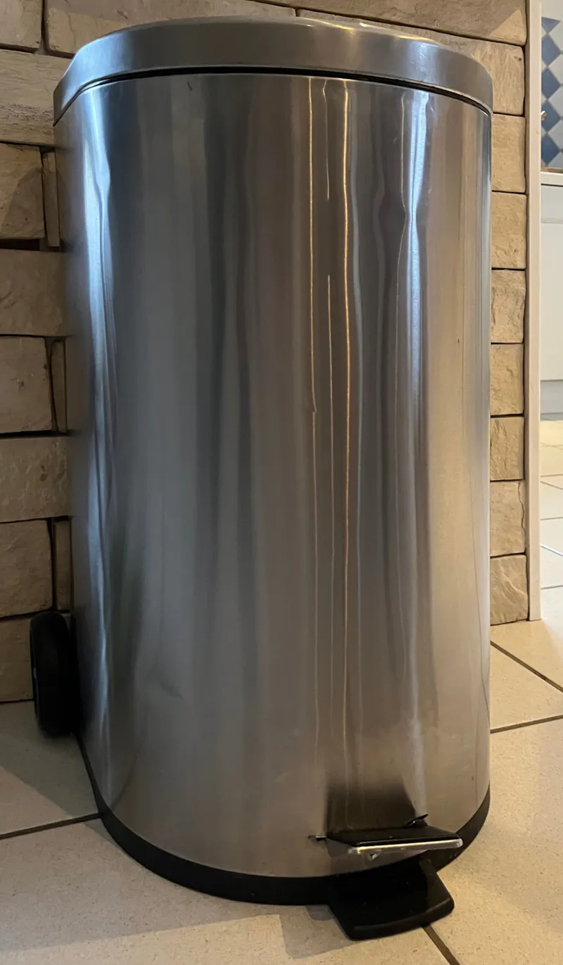 Poubelle à pédale en inox posée sur un carrelage clair, contre un mur en pierres claires