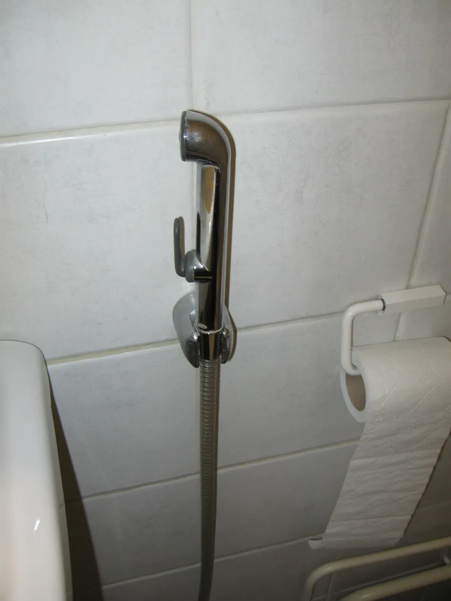 Douchette WC à main en métal accrochée au mur carrelé blanc, à côté d'un rouleau de papier toilette